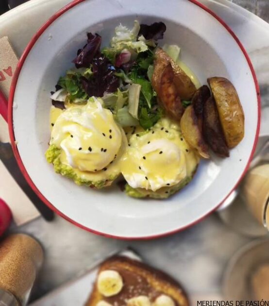 Un plato de huevos Benedict con aguacate y ensalada, acompañado de patatas asadas, servido en una mesa.