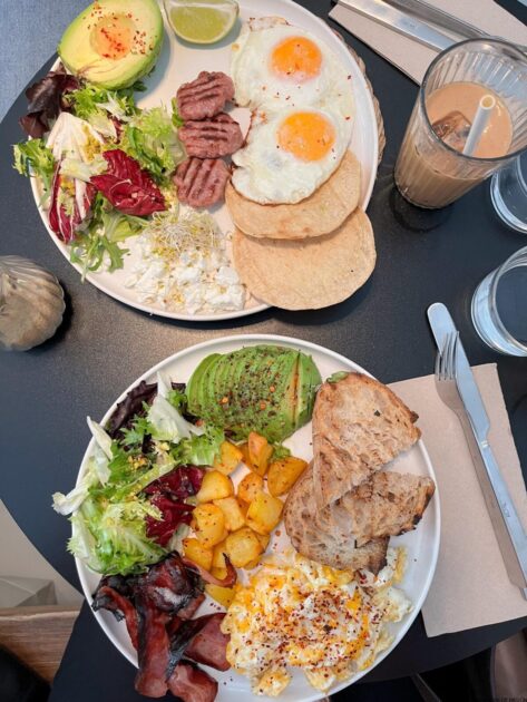Dos platos de desayuno con huevos, tocino, aguacate, ensalada y pan junto con tazas de café y un batido sobre una mesa gris.