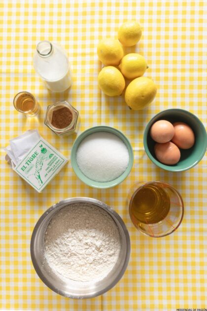 Sobre un mantel de cuadros amarillos están dispuestos varios ingredientes para hornear, entre ellos harina, azúcar, huevos, limones, leche, canela, miel y una botella de mantequilla con la etiqueta "El Tigre".