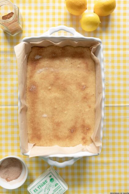 Un pastel rectangular en una fuente para hornear blanca con papel pergamino, colocado sobre un mantel a cuadros amarillo y blanco. Sobre la mesa se ven limones y un recipiente de canela junto con un cuenco de azúcar y canela.