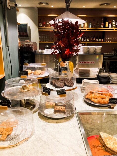 El mostrador de una panadería que muestra varios pasteles bajo cubiertas transparentes con etiquetas, junto a una máquina de café expreso y tazas apiladas.
