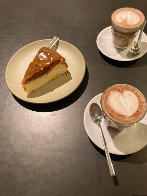 Una rebanada de tarta de queso con caramelo en un plato con un tenedor y dos cafés con leche con espuma en forma de corazón sobre una mesa oscura.