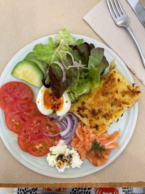 Un plato de comida con una rodaja de frittata, salmón ahumado, ensalada fresca, rodajas de aguacate, tomates, un huevo cocido y un tenedor a un lado.