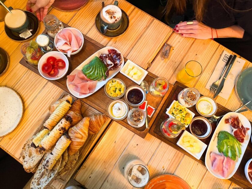 Vista superior de una mesa de desayuno con varios platos, incluidos croissants, embutidos, quesos y bebidas, con las manos de una mujer visibles.