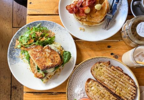 Vista aérea de un desayuno extendido sobre una mesa de madera, que incluye panqueques con tocino, un sándwich de huevo y pan tostado con una guarnición de salsa.