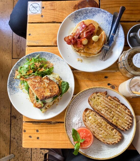 Vista aérea de un desayuno extendido sobre una mesa de madera, que incluye panqueques con tocino, un sándwich de huevo y pan tostado con una guarnición de salsa.