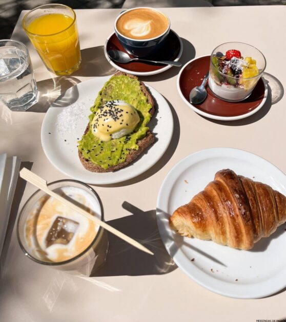 Mesa de desayuno con tostadas de aguacate, croissant, un plato de fruta y tres bebidas, incluido café y jugo de naranja, iluminada por la luz del sol.