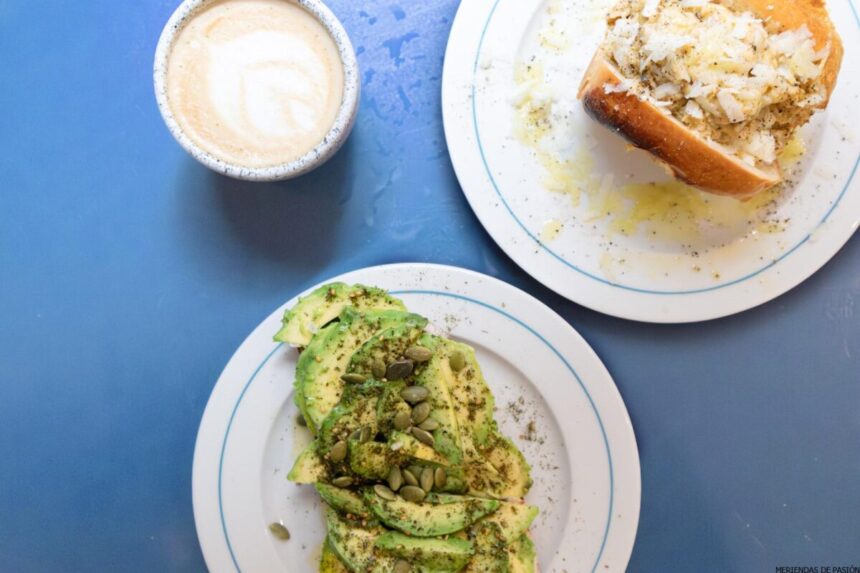 Una vista superior de una comida que consiste en un plato con aguacates en rodajas cubiertos con hierbas y semillas, otro plato con pan y queso y una taza de café con latte art sobre una superficie azul.