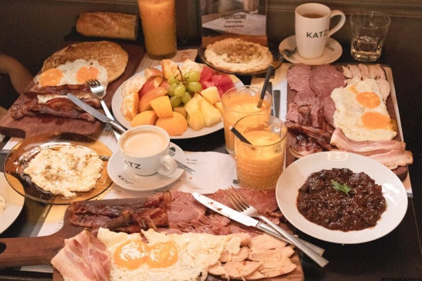 Hay una mesa puesta con varios alimentos para el desayuno, incluidos huevos, jamón, tocino, fruta fresca, panqueques, café y jugo de naranja.