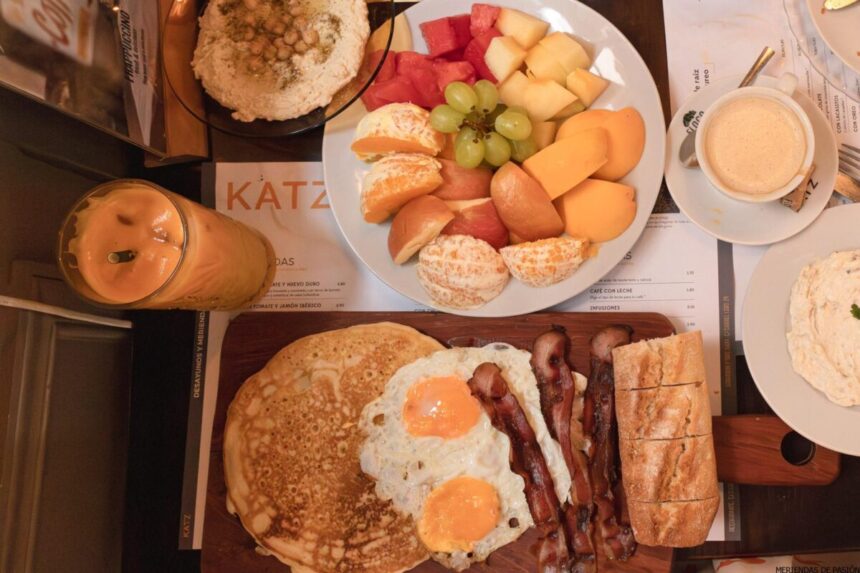 Un desayuno acompañado de panqueques, huevos fritos, tocino, pan, frutas variadas, un plato de hummus, un vaso de jugo de naranja y una taza de café sobre una mesa.