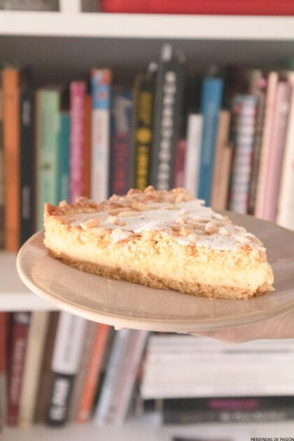 Se coloca una rebanada de tarta de almendras sobre un plato de madera, sobre el fondo de una estantería llena de varios libros.