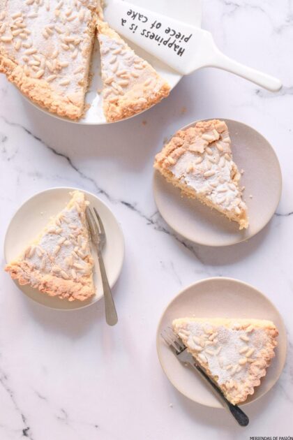 Tres rebanadas de pastel cubierto de almendras en platos blancos, con una rebanada en un servidor de pastel que dice "La felicidad es pan comido" sobre una superficie de mármol. Se colocan dos tenedores al lado de las rodajas.