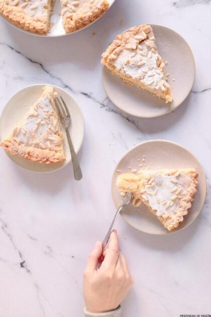 Tres rebanadas de pastel de almendras en platos con tenedores, una rebanada sostenida con una mano sobre una superficie de mármol.