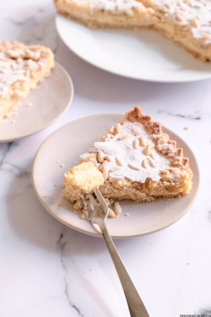 Un trozo de pastel cubierto de migas con un tenedor sobre un plato blanco. El pastel se adorna con azúcar en polvo y piñones, con porciones de pastel adicionales al fondo.