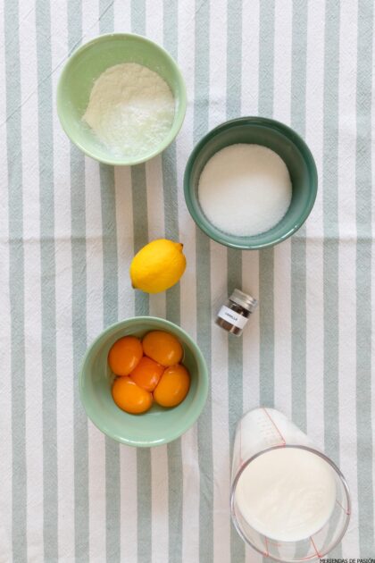 Ingredientes para una receta sobre un mantel rayado: un cuenco de harina, un cuenco de azúcar, un limón, un recipiente pequeño de vainilla, un cuenco con yemas de huevo y un vaso medidor con leche.