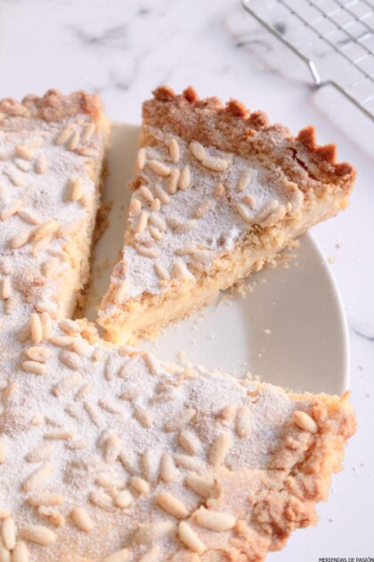 Un plato con una tarta de almendras en rodajas cubierta con azúcar glass y piñones.