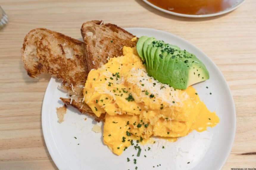 Un plato blanco con huevos revueltos, aguacate en rodajas y dos trozos de pan tostado, aderezado con queso rallado y hierbas picadas.
