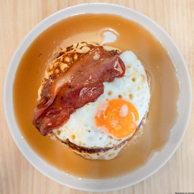 Un plato de desayuno que incluye una pila de panqueques cubiertos con un huevo frito, tocino crujiente y rociados con almíbar, servido en un plato blanco.