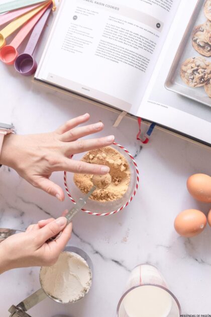 Una persona mide el azúcar moreno con una cucharadita sobre un tazón, con un libro de cocina abierto, cucharas medidoras, harina, huevos y un vaso de leche cerca en la encimera de la cocina.