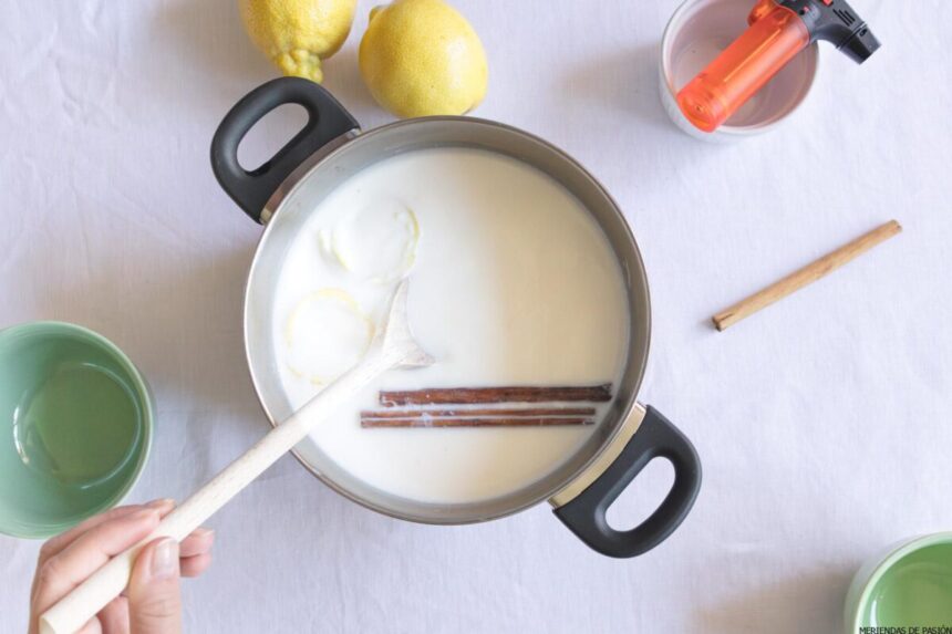 Persona revolviendo leche con palitos de canela y cáscaras de limón en una olla. Alrededor de la olla hay limones, un cuenco verde, un cuenco blanco y un soplete de cocina sobre una superficie blanca.