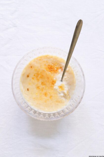 Un tazón de arroz con leche cremoso cubierto con una pizca de canela o azúcar, con una cuchara de plata apoyada en él, colocado sobre una superficie blanca.