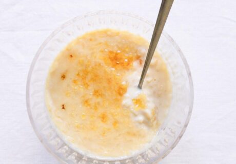 Un tazón de arroz con leche cremoso cubierto con una pizca de canela o azúcar, con una cuchara de plata apoyada en él, colocado sobre una superficie blanca.