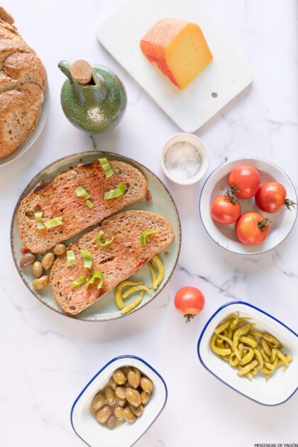 Un plato con rebanadas de pan coronadas con tomates y pimientos verdes, rodeado de tomates, queso, aceitunas verdes, pimientos y sal sobre una superficie de mármol.