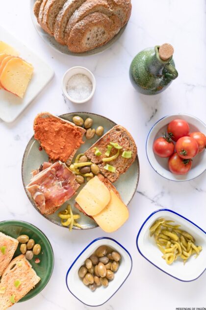 Una mesa con pan, quesos, aceitunas, tomates ramillet, judías verdes y una pequeña botella de aceite, dispuestos en platos blancos y verdes.