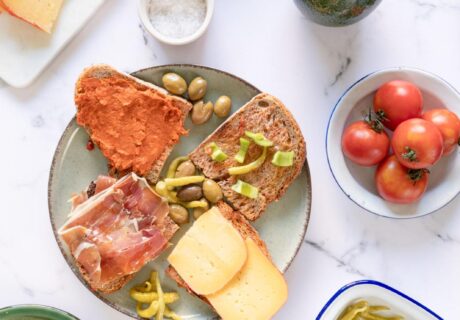 Una mesa con pan, quesos, aceitunas, tomates ramillet, judías verdes y una pequeña botella de aceite, dispuestos en platos blancos y verdes.