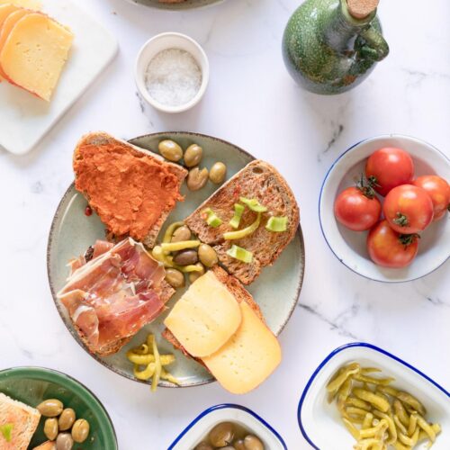 Una mesa con pan, quesos, aceitunas, tomates ramillet, judías verdes y una pequeña botella de aceite, dispuestos en platos blancos y verdes.