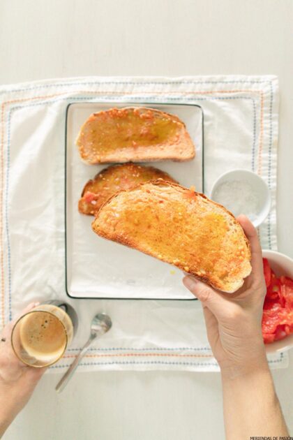 Mano que sostiene una rebanada de pan tostado con tomate encima de un plato con más pan tostado. Café, tomates y sal visibles en el costado.